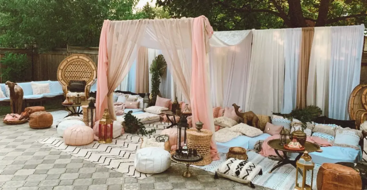 Bohemian-style outdoor setup for a henna event, featuring sheer draped canopy, pillows, poufs, and rugs in soft pastel shades. Decorative items include lanterns, a camel figurine, and intricate wicker furniture, all set on a patterned patio beneath trees.