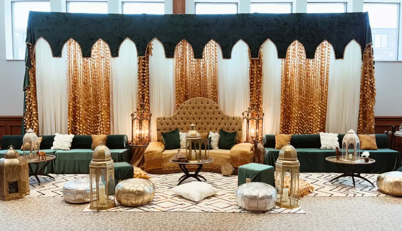 A lavishly decorated room features a green and gold theme with a large ornate backdrop. Green and gold sofas surround a grand tufted chair. Elegant lanterns, cushions, and low tables with intricate patterns enhance the luxurious ambiance, perfect for henna event decor.