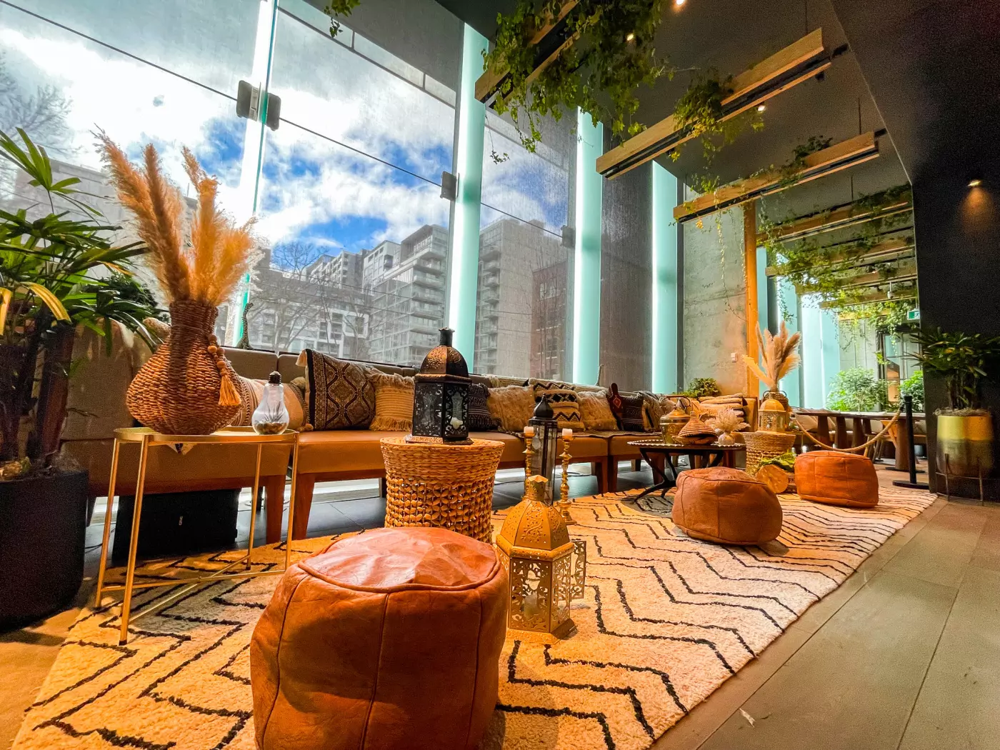 A chic lounge with a boho-inspired decor featuring leather poufs, wicker baskets, and a patterned rug. Large windows let in ample natural light. Potted plants and hanging greenery add a natural touch, while pillows and lanterns enhance the cozy atmosphere.
