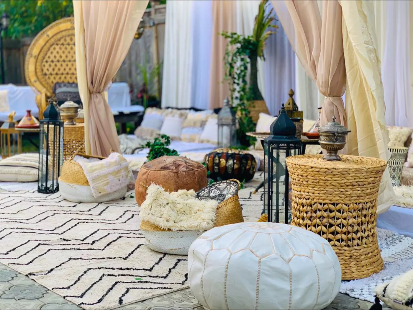 A cozy outdoor lounge area is decorated with white and brown poufs, woven baskets, and large pillows on a patterned rug. Draped beige curtains frame the space, while lanterns and lush greenery enhance the serene, inviting ambiance befitting Moroccan themed events decor.