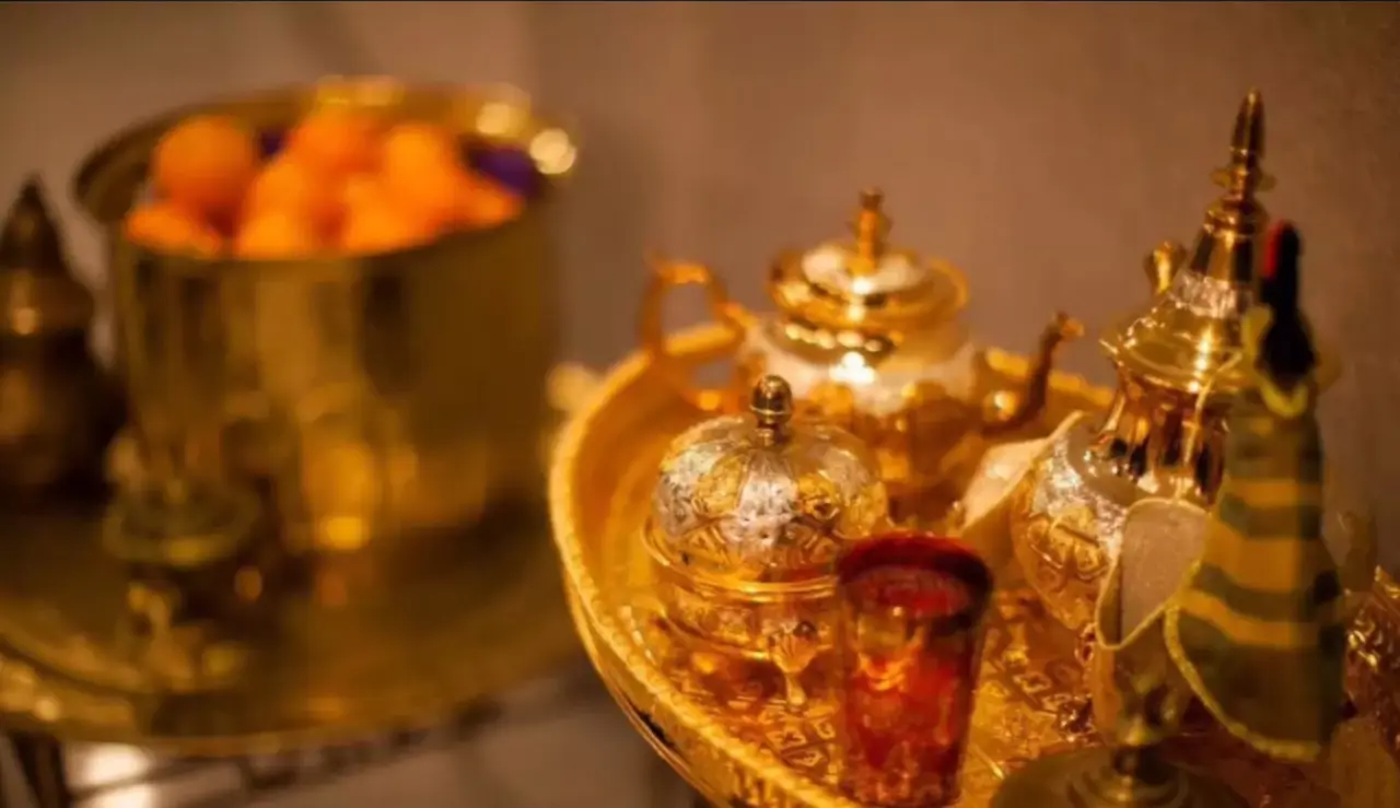 A golden tray holds intricately designed teapots, a red glass, and other ornate items, perfect for Moroccan decor rentals. In the background, a golden bowl filled with orange fruits is visible. The scene appears to be set in a warm, softly lit environment, highlighting the luxurious metallic props.